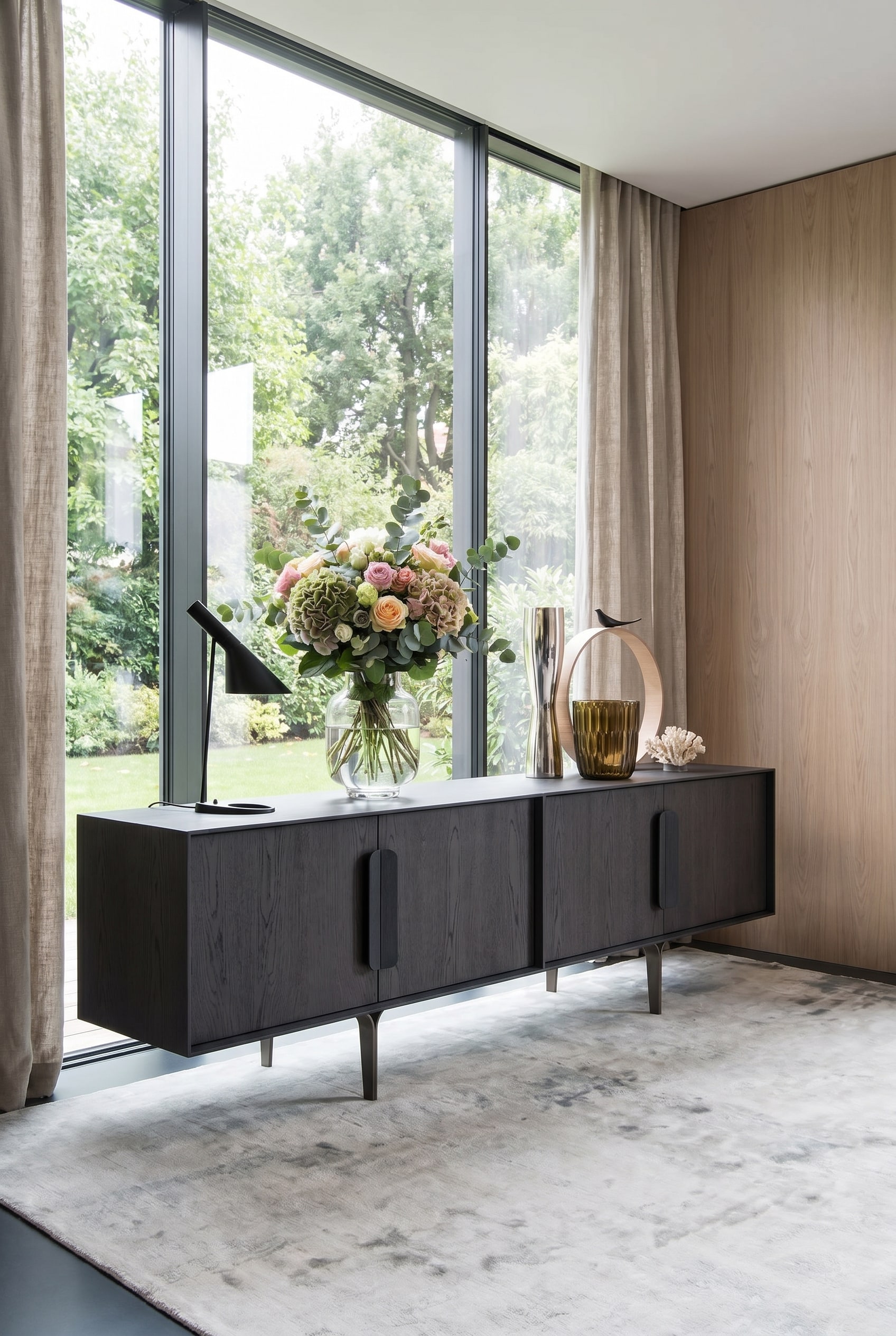Dunkles Designer-Sideboard vor großen Fenstern, darauf eine helle, leuchtende Statement-Vase mit Frühlingsblumen Dunkles Designer-Sideboard vor großen Fenstern, darauf eine helle, leuchtende Statement-Vase mit Frühlingsblumen