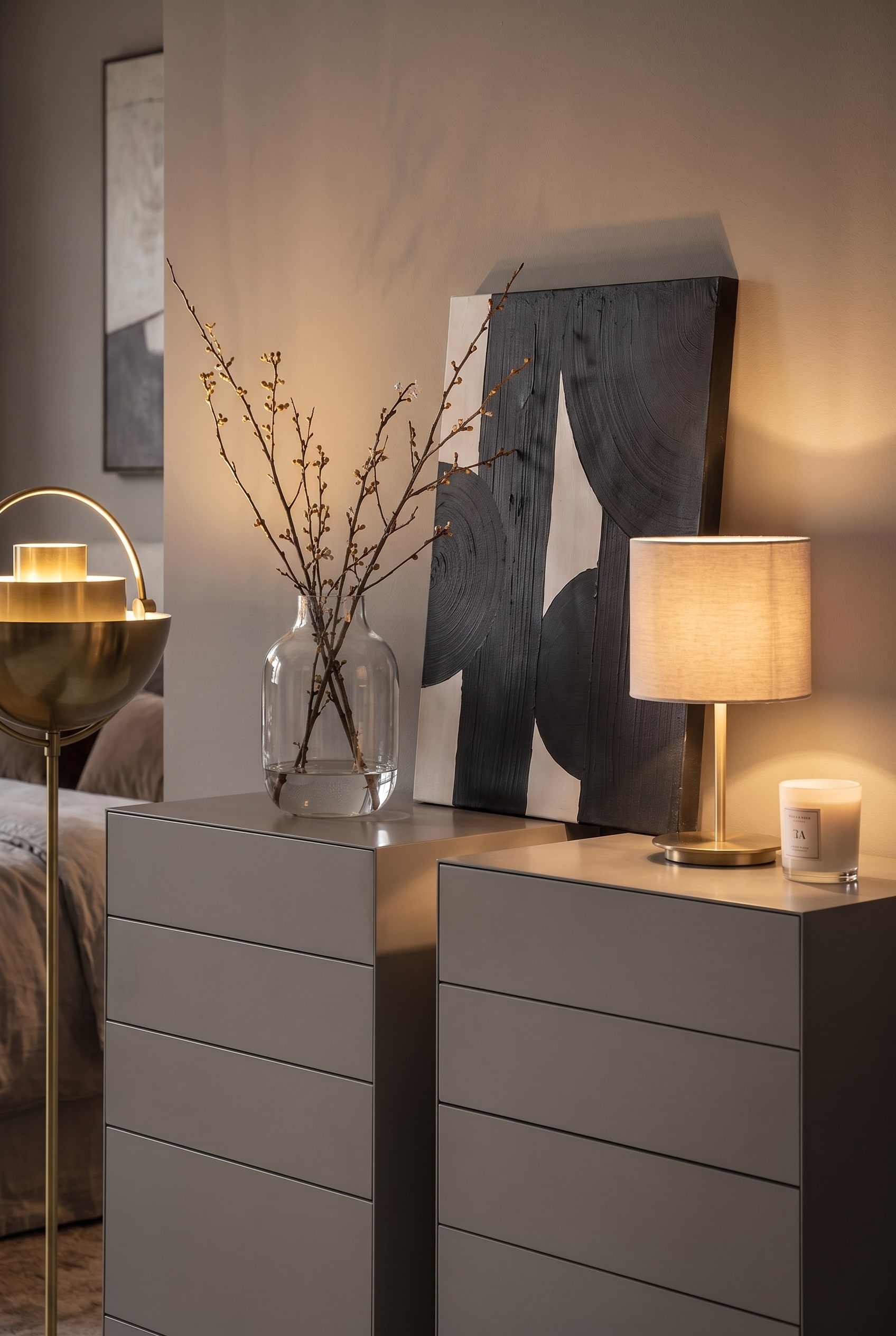 Decoration on two gray tall chests of drawers: A leaning abstract picture, a glass vase with branches, and a table lamp.