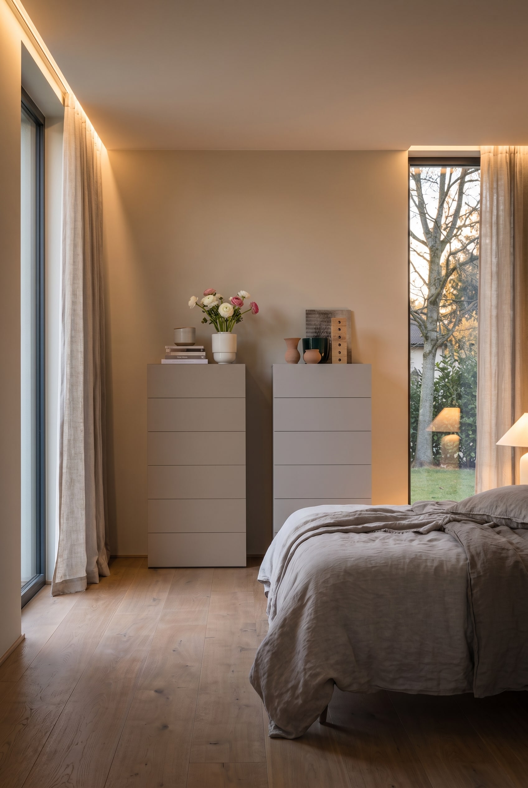 Two greige tall chests of drawers in a modern bedroom with linen bedding and floor-to-ceiling windows.