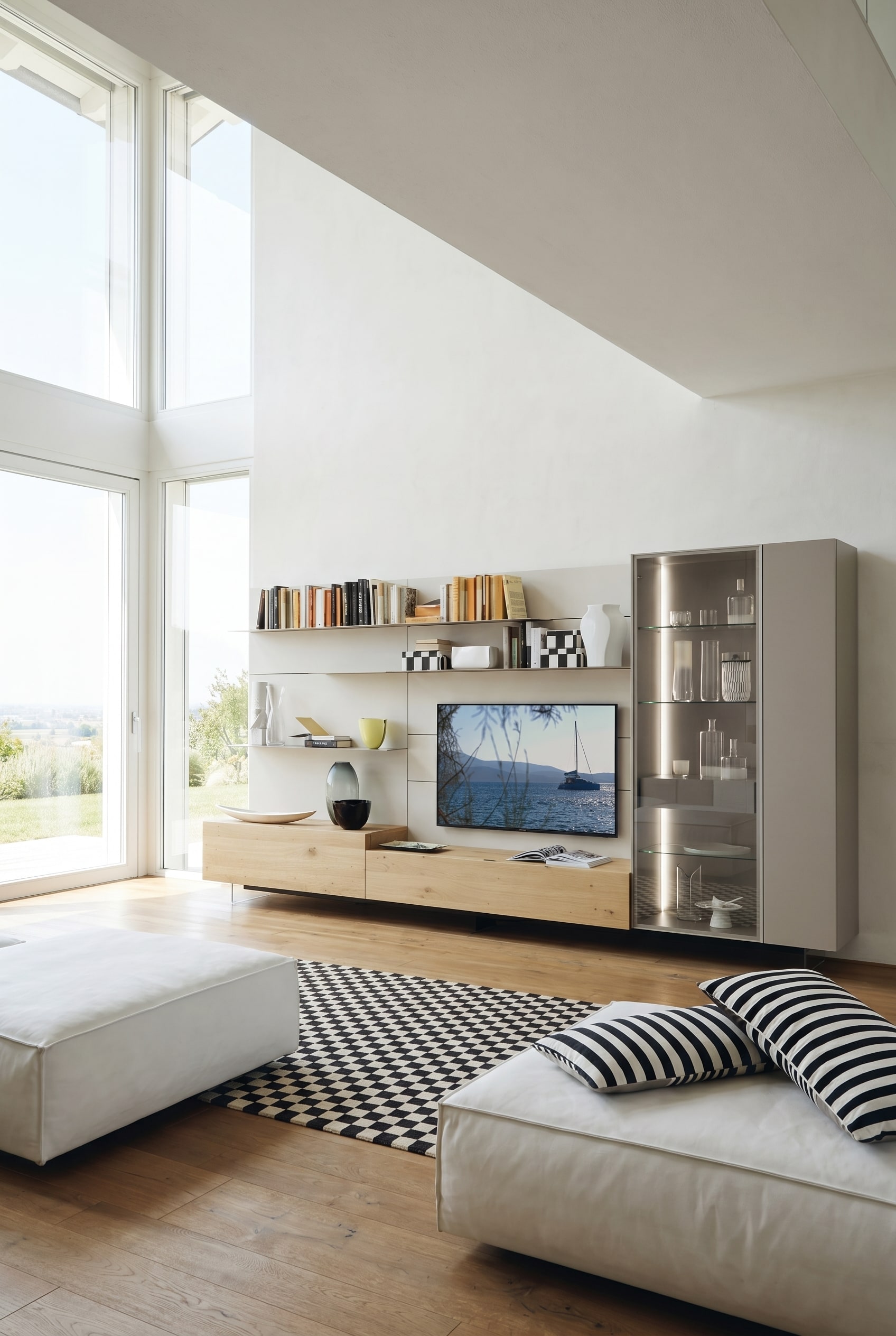 Helles Wohnzimmer mit schwebenden Basismodulen aus heller Eiche, wei&szlig;en Regalb&ouml;den und einer hohen, warm beleuchteten Glasvitrine