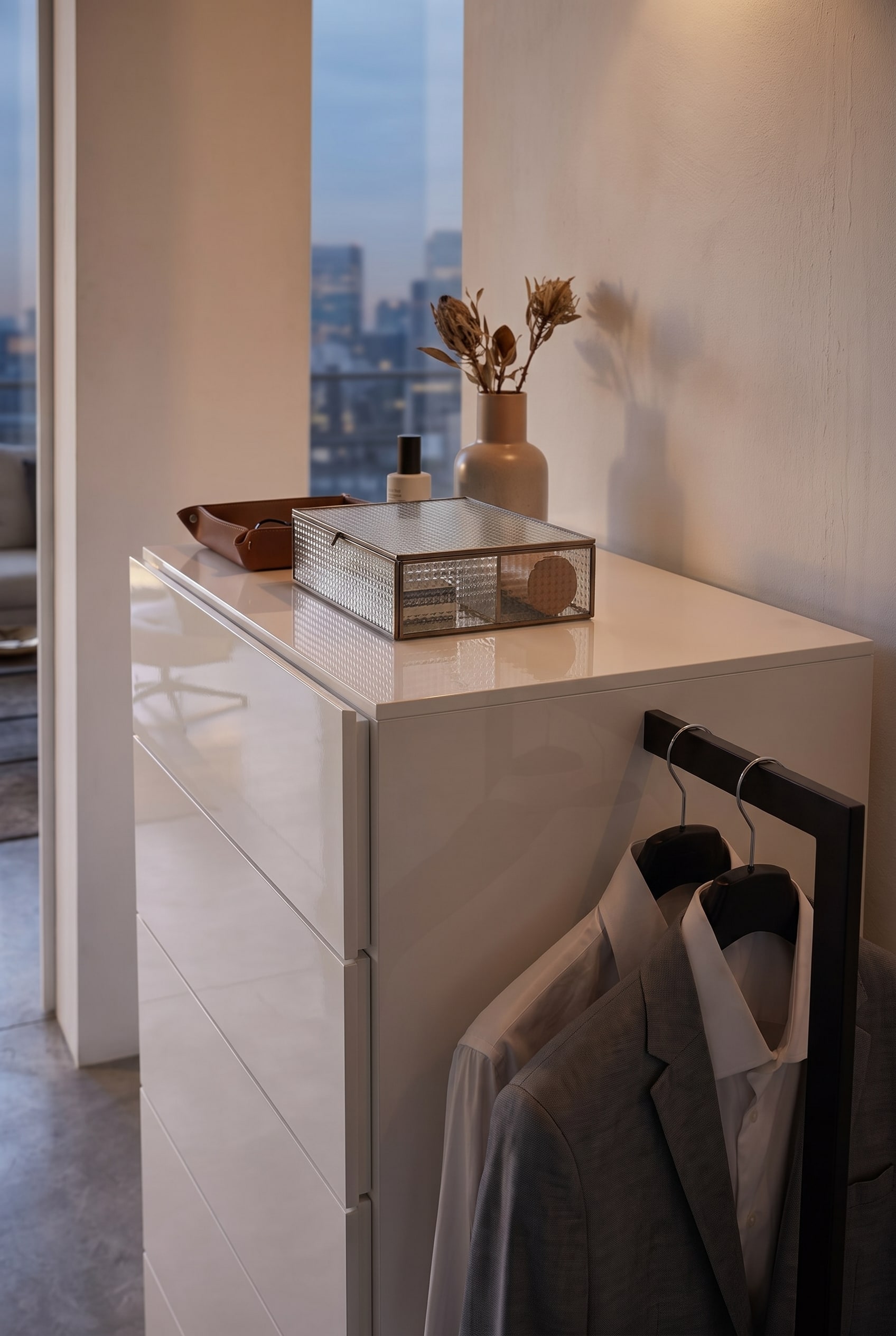Detail shot of a white chest of drawers with a ribbed glass box, leather tray, and a side clothes rail with a grey blazer.