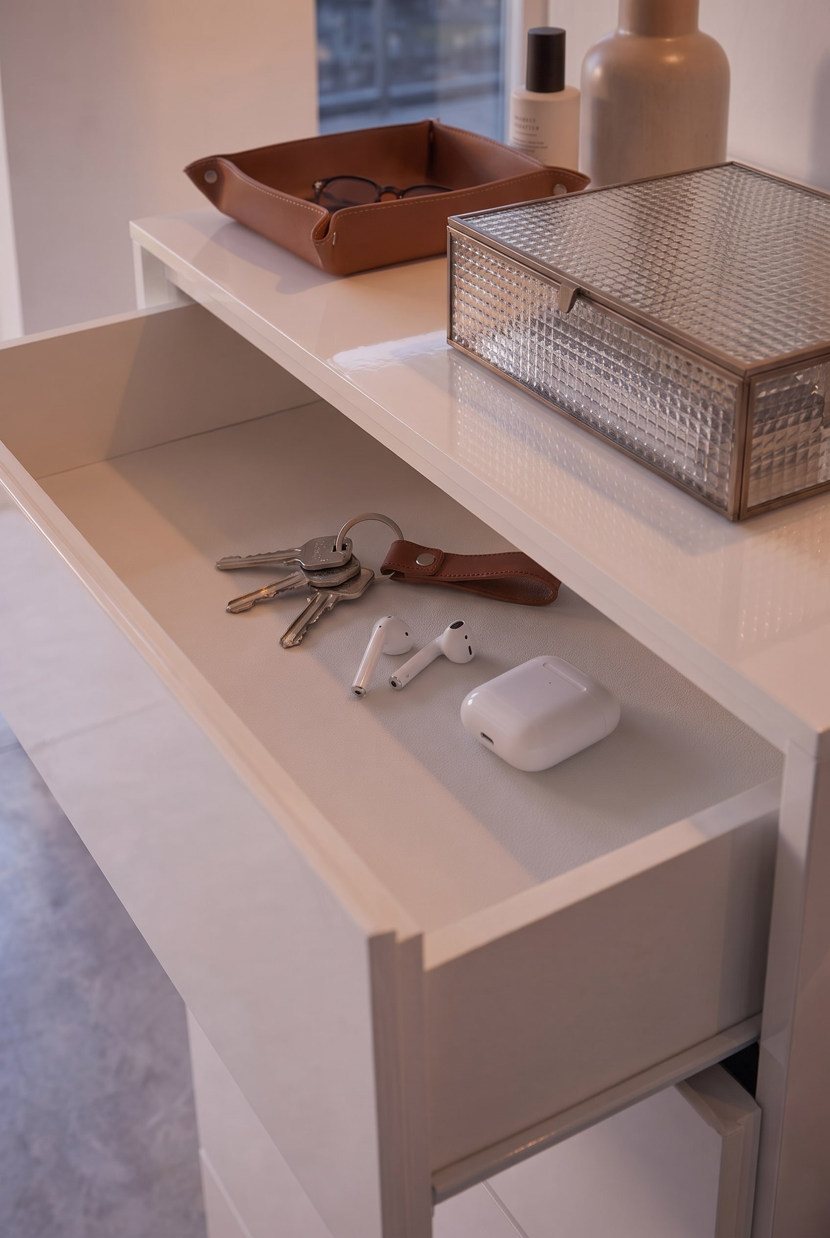 Open shallow drawer of a white dresser, with keys, AirPods, and a leather tag neatly arranged inside.