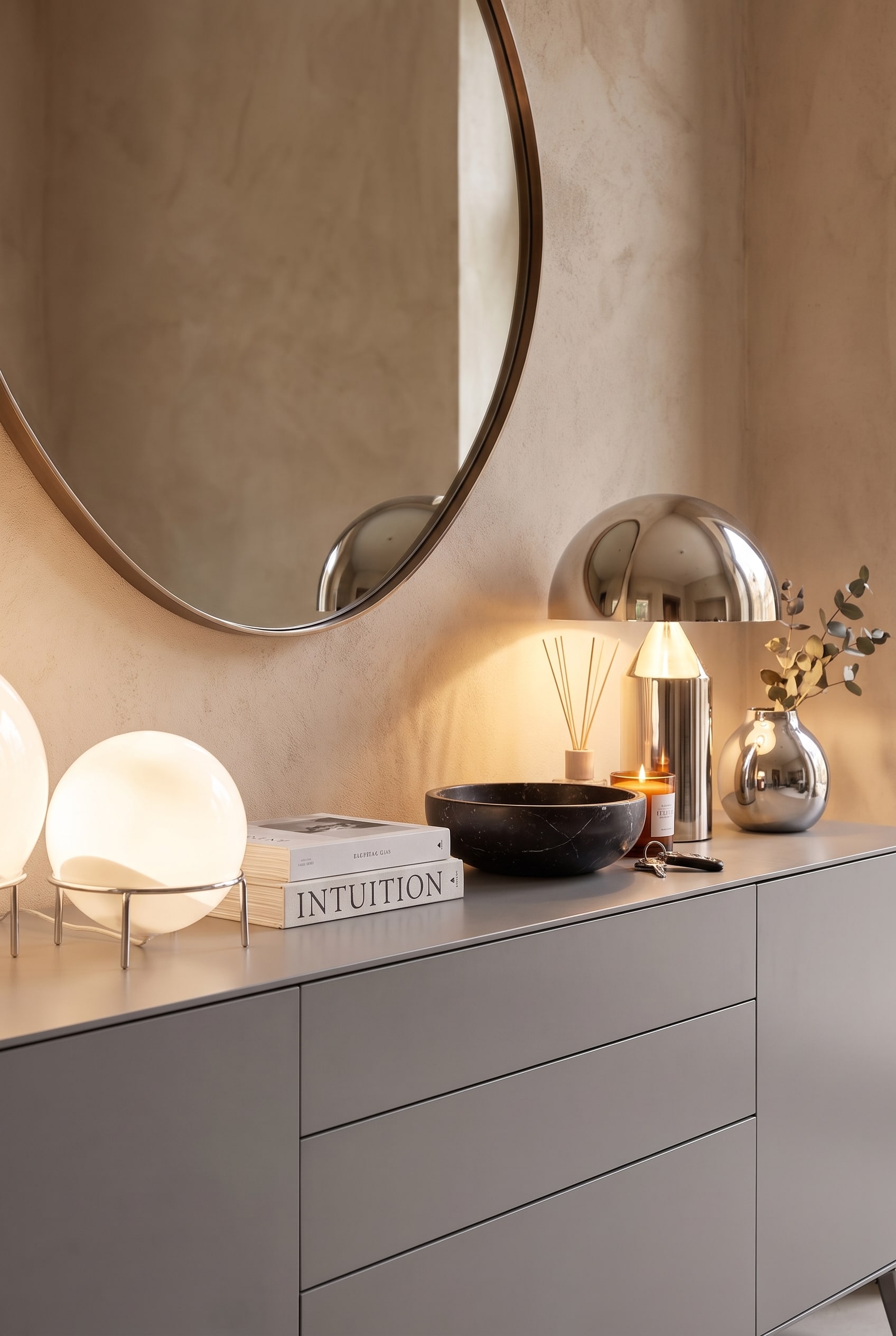 Detailaufnahme der Sideboard-Dekoration mit zwei runden, leuchtenden Tischlampen, einer schwarzen Marmorschale und Büchern auf matter Oberfläche.