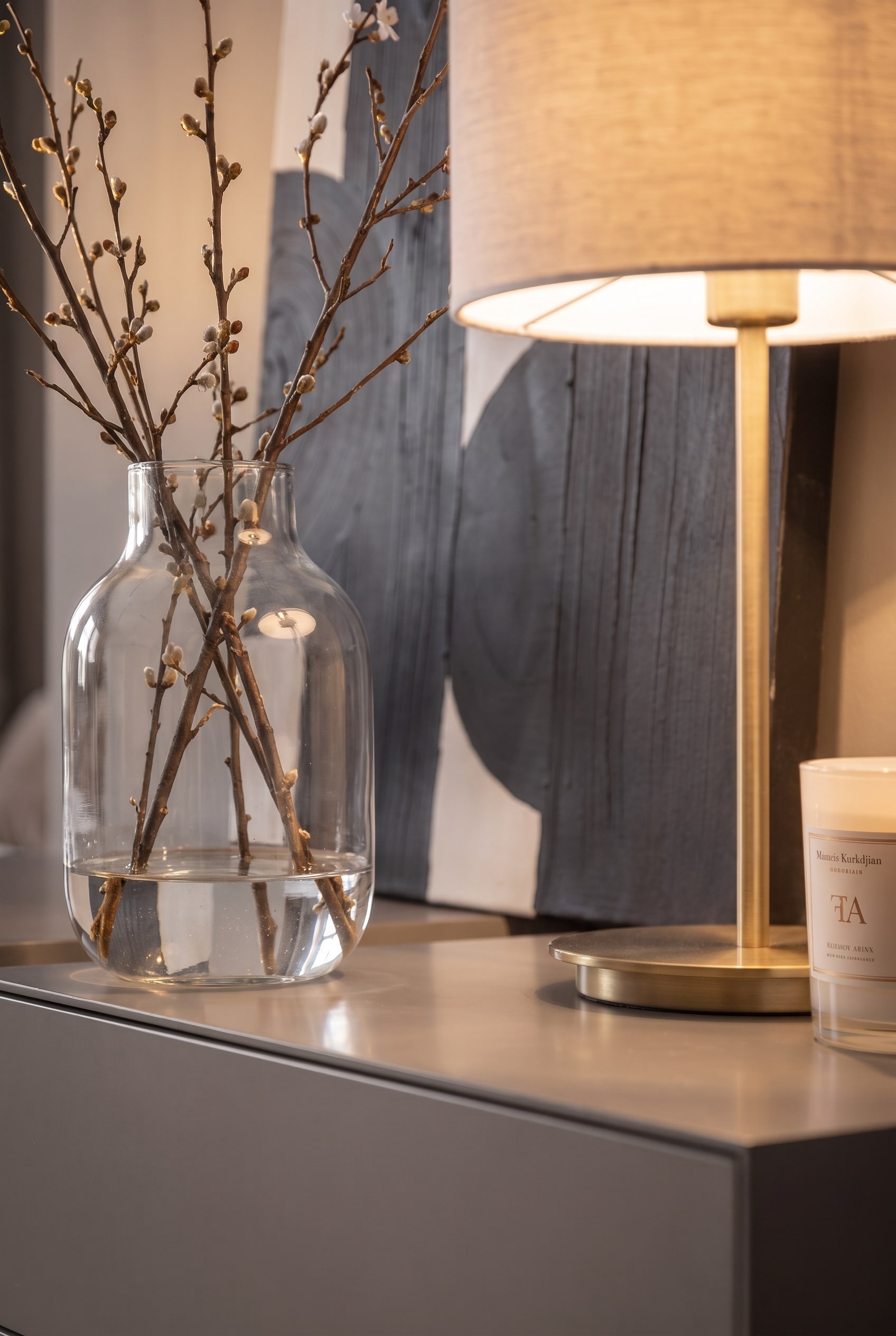 Detailed view of a bulbous glass vase with branches next to a lit table lamp and a candle on a chest of drawers.