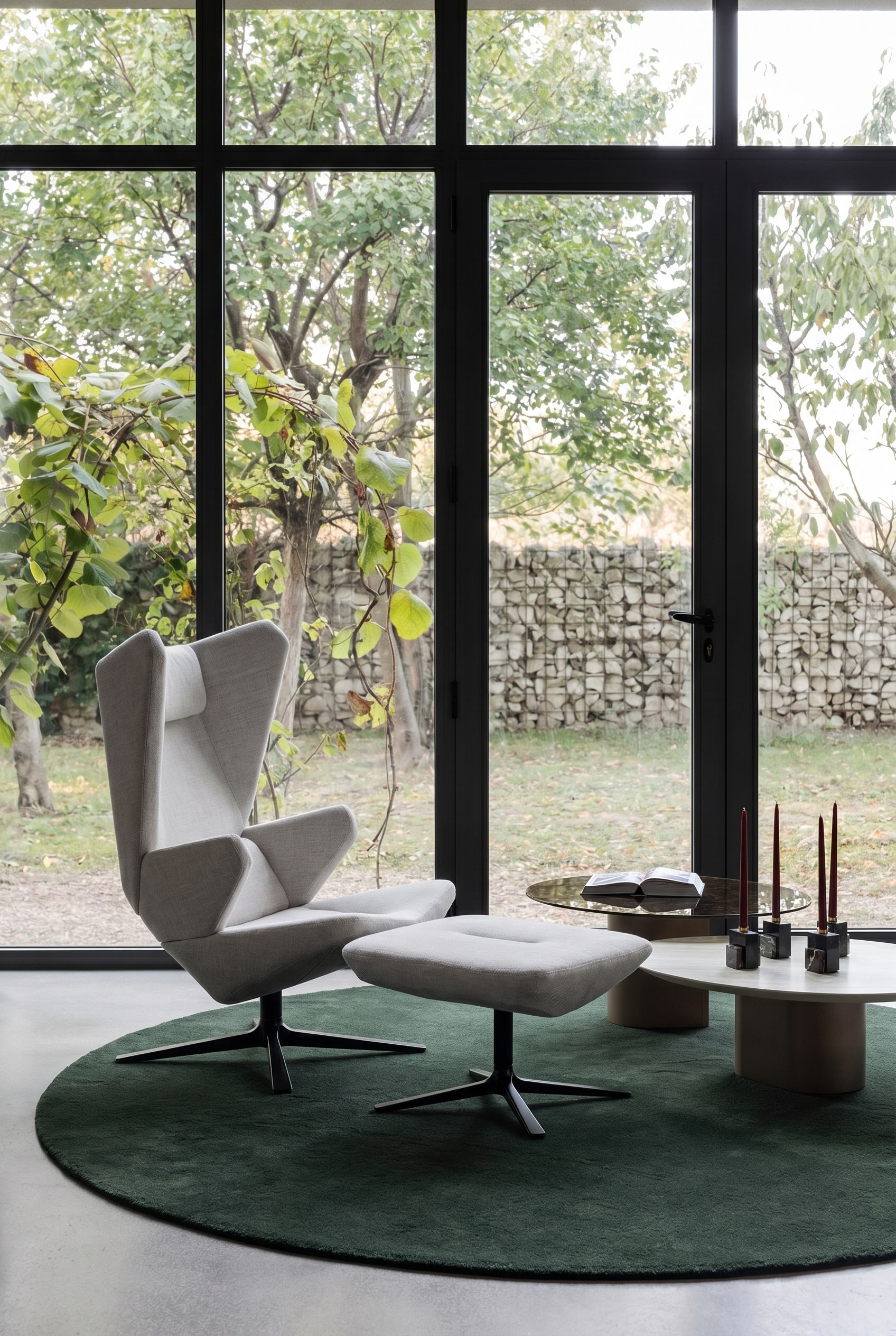 Skulpturaler grauer Sessel mit hohem R&uuml;cken auf einem dunkelgr&uuml;nen runden Teppich in einem minimalistischen Wohnzimmer mit gro&szlig;en Fenstern